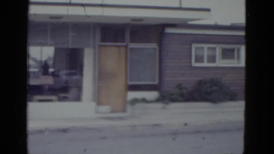 1952 New Orleans commercial building mid-pan on 8mm; plate-glass window with reflected period car, wooden door, and horizontal reddish-brown siding.