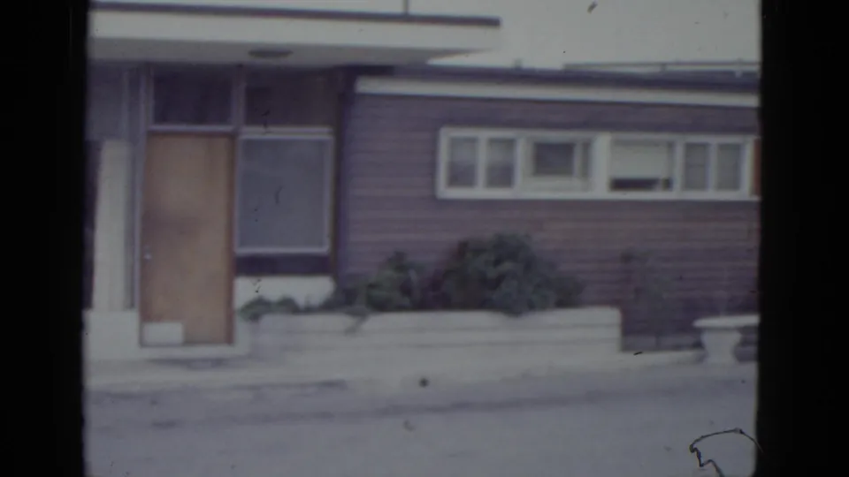 8mm pan of a 1950s New Orleans commercial building; wooden door at frame edge, white-framed louvered windows, horizontal siding, and low concrete planter with shrubbery.