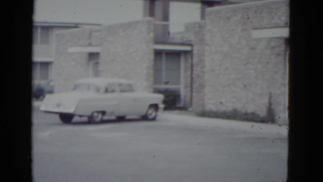 Large American sedan of early 1950s styling parked before a rough stone-clad mid-century building, 8mm film circa 1952.
