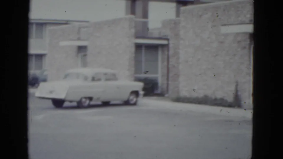 Large American sedan of early 1950s styling parked before a rough stone-clad mid-century building, 8mm film circa 1952.