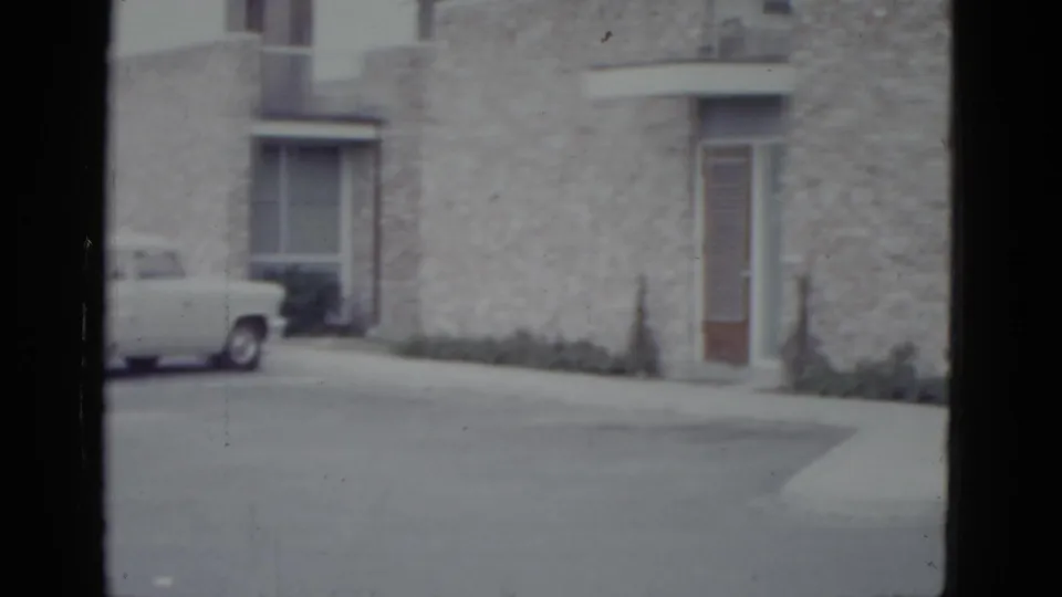 Rough stone building facade with wooden entrance door and low shrubs; partial view of 1950s sedan at frame left, 8mm circa 1952.