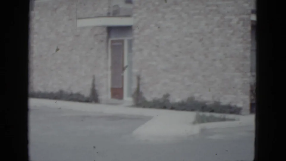 Rough stone building facade with wooden entrance door and low shrubs, camera continued pan right, no vehicle visible, 8mm circa 1952.