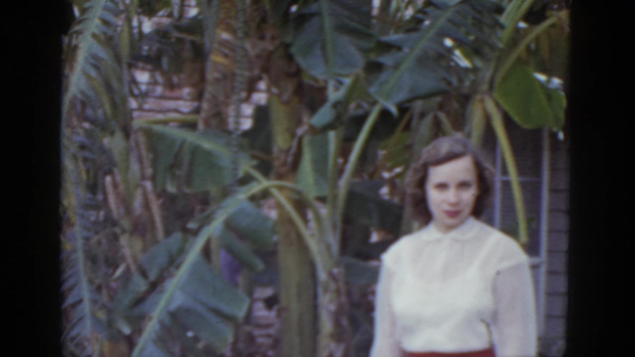 Woman in white blouse and red waistband facing toward camera, surrounded by large-leaved banana plants.
