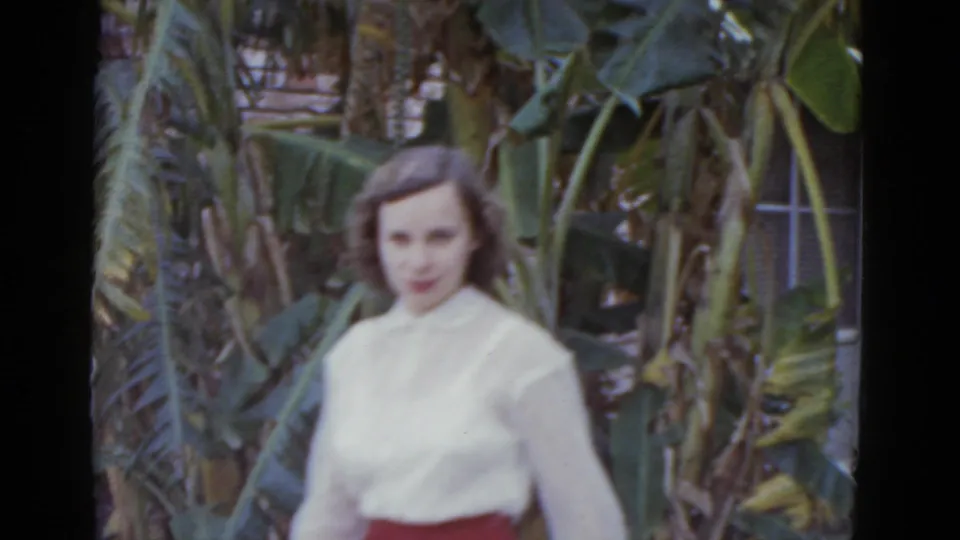 Young woman in white blouse and red skirt standing in garden; banana trees with a fruit cluster behind her; 1952 8mm footage, New Orleans.