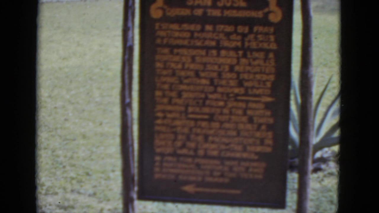 Brown wooden sign with orange text panels on two wooden posts, green lawn in background, agave plant visible at right.