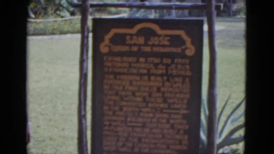 San José Mission sign between two wooden posts; agave plant foreground right; green mowed lawn; 8mm color, circa 1952.