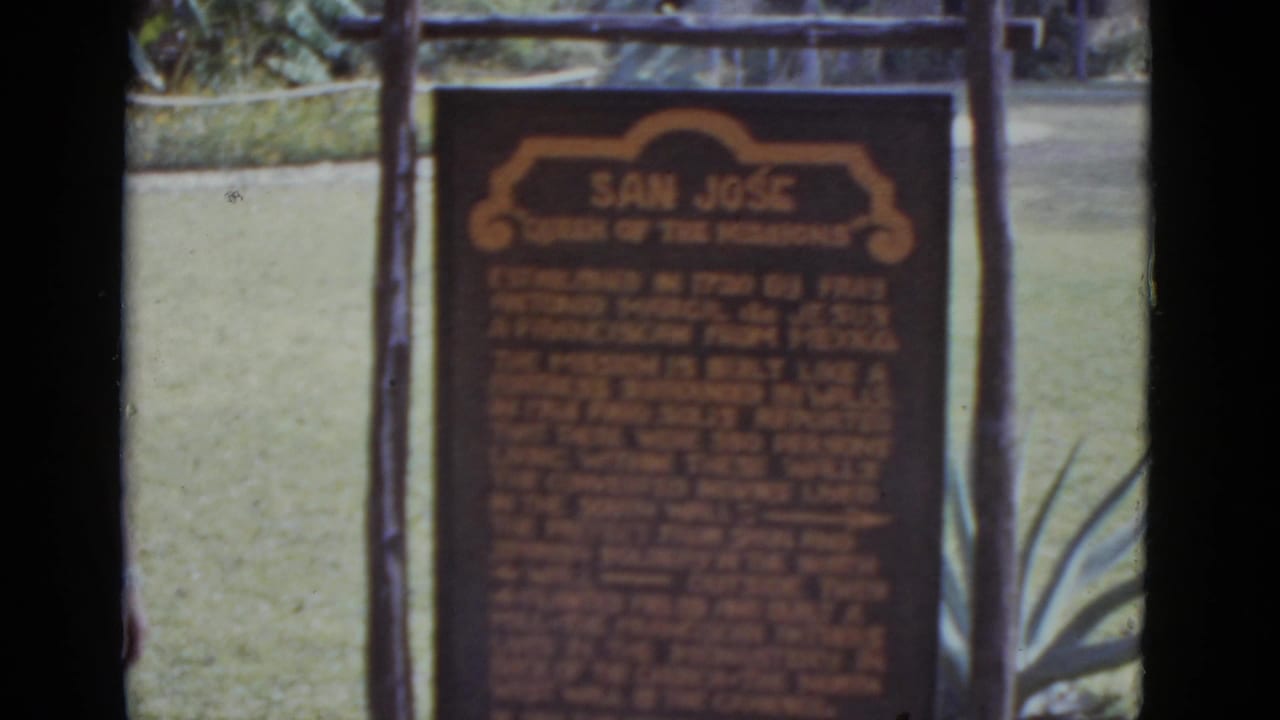 Dark brown mission plaque reading 'San Jose Queen of the Missions,' wooden post frame at left, agave plants at right, sandy forecourt