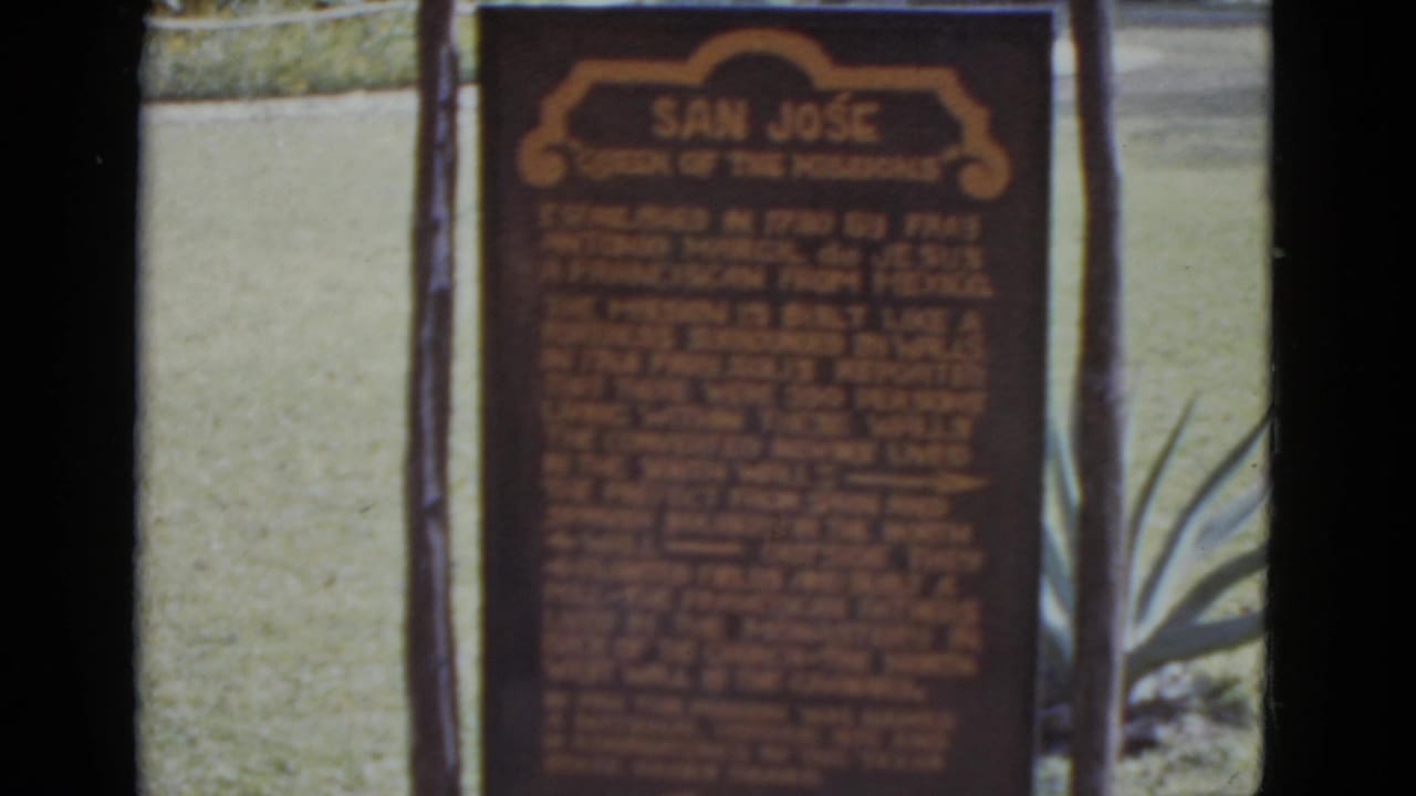 1952 8mm home movie close-up of the 'San Jose Queen of the Missions' historical marker centered in frame; wooden posts and agave foliage flank the sign