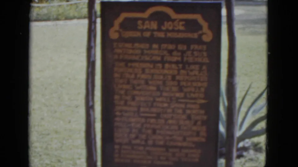 1952 8mm home movie close-up of the 'San Jose Queen of the Missions' historical marker centered in frame; wooden posts and agave foliage flank the sign