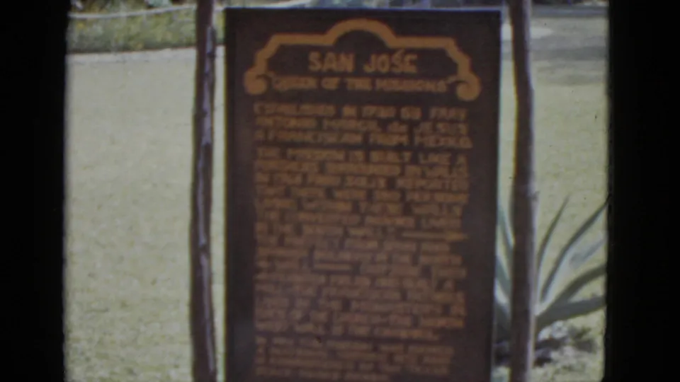 Mid-clip 8mm frame c.1952 showing the 'San Jose Queen of the Missions' historical marker; wooden posts and agave plants frame the dark brown sign