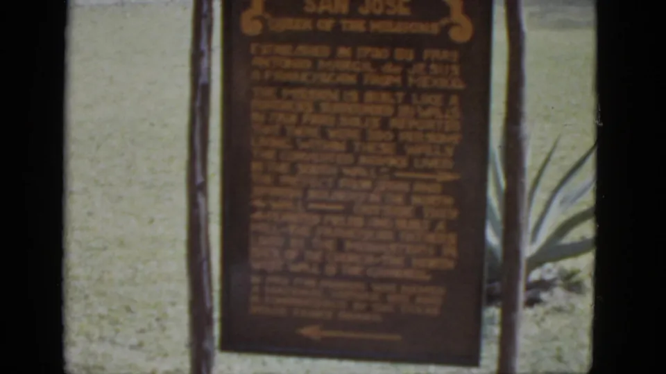 Closing 8mm frame c.1952 of the 'San Jose Queen of the Missions' historical marker; dark wooden posts flank the plaque with agave foliage at right