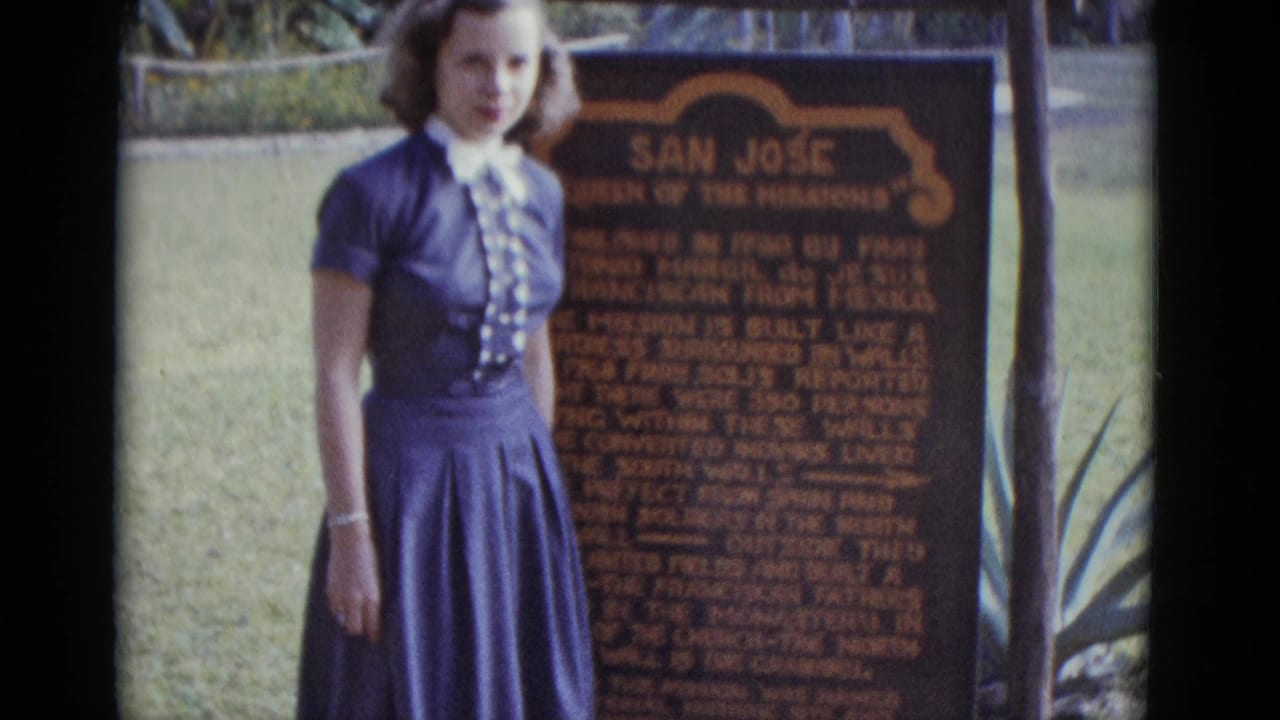 Young woman in blue dress standing beside a wooden 'San José — Queen of the Missions' historical marker on a grassy lawn.