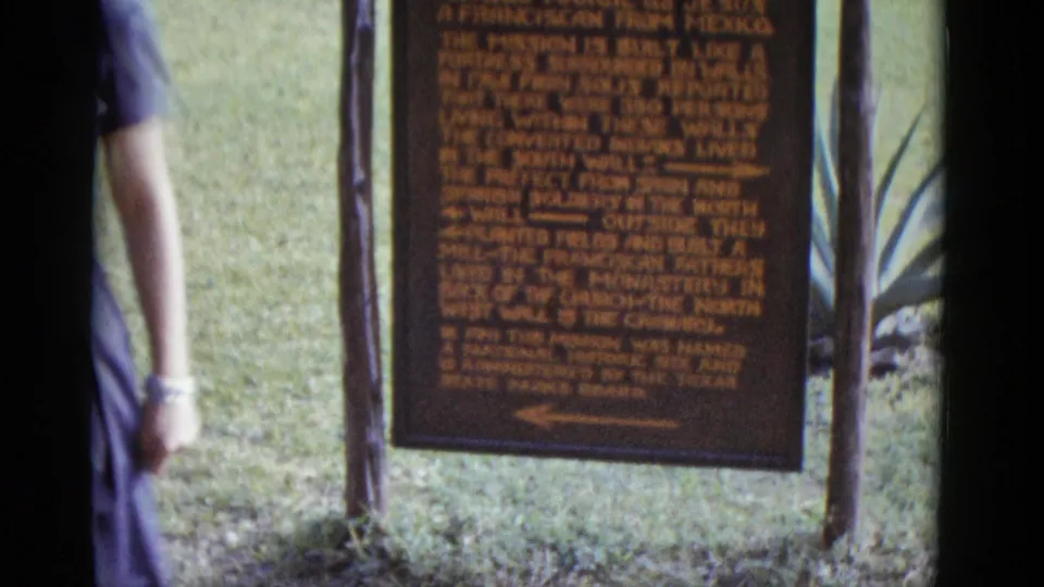 Partial figure of a woman in a 1950s blue dress near the Mission San José historical marker, showing natural handheld camera movement, circa 1952.