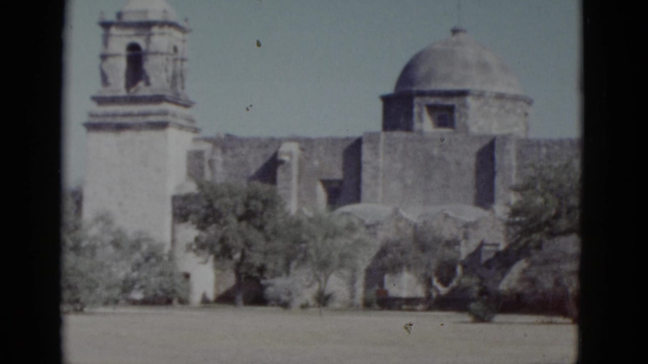 White stone mission-style building with bell tower and dome, framed by sparse scrub trees against a pale sky.