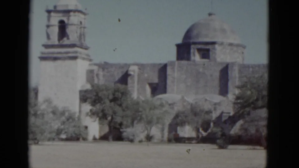 Wide view of a Spanish Colonial mission-style structure showing a bell tower and rounded dome with desert scrub trees in foreground — 8mm, likely American Southwest, 1952.