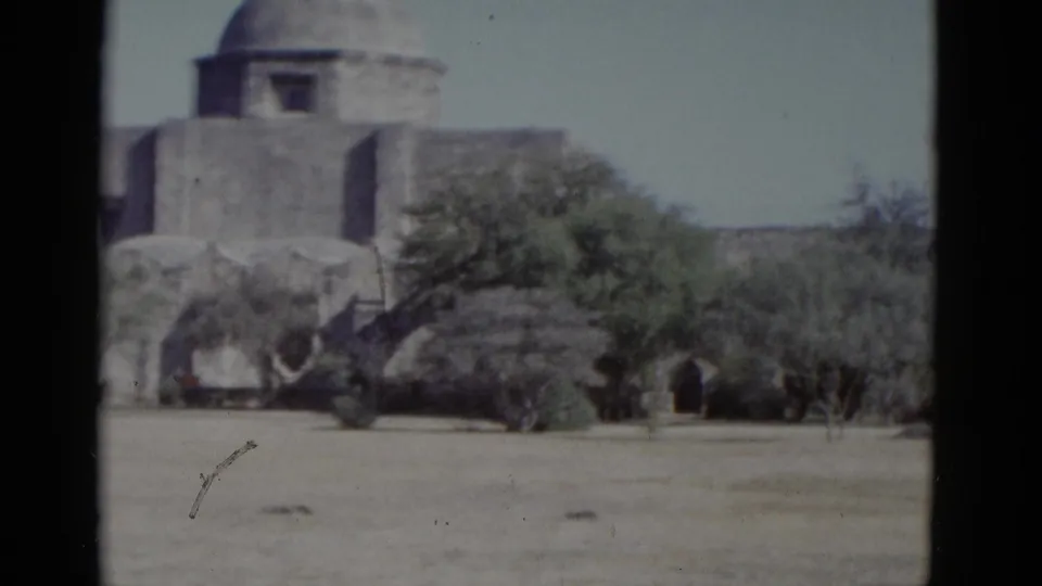 Exterior of a mission-style building with dome framed by desert scrub — 1952 8mm footage with a visible film scratch in the lower frame area.