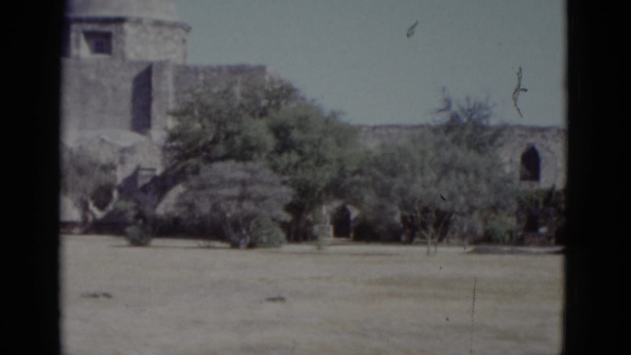Stone mission-style arcade and corner tower viewed across a dry sandy field, green trees and pale sky, 8mm film.