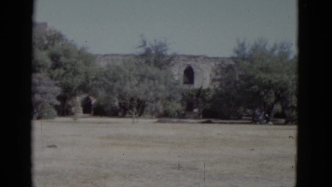 Two arched openings in a low stone wall seen across an arid field with green trees, 8mm home movie.