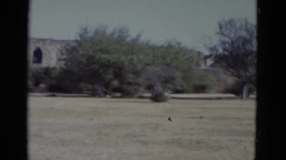 1952 8mm home movie establishing shot: arid open field, sparse trees, and a stone structure with an arched window, possibly at a Southwest US historic site.