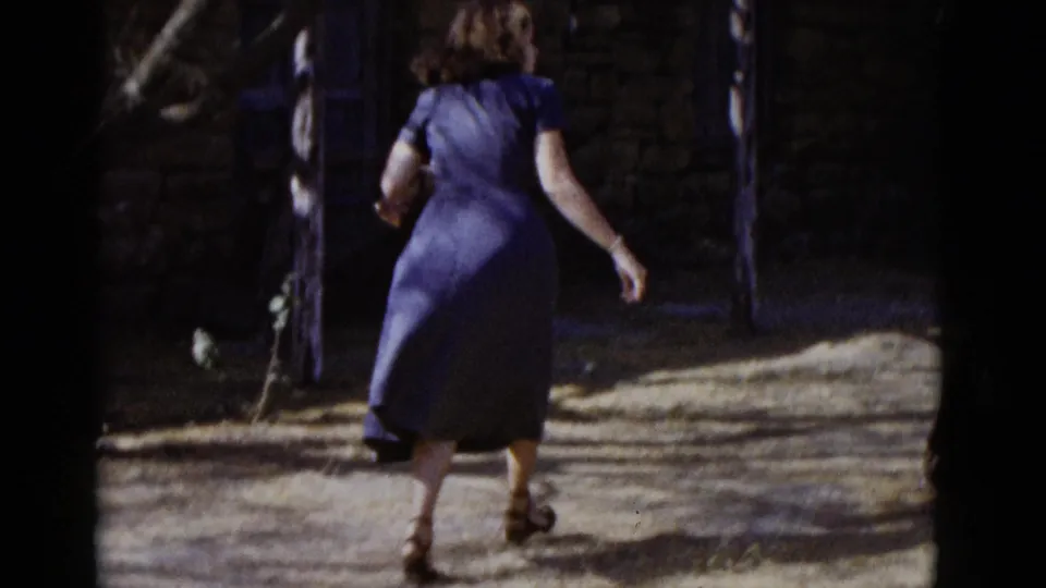 1952 8mm home movie: woman in a blue short-sleeve dress and dark heeled shoes walks away from camera past a stone wall and wooden post.
