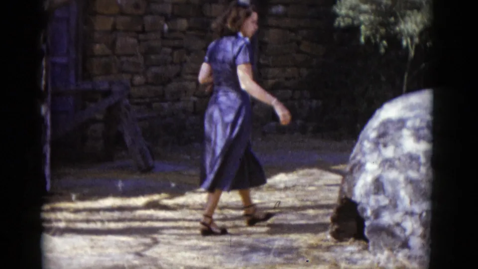 1952 8mm home movie: woman in blue dress in three-quarter profile walking past a stone masonry wall with a blue wooden gate; arid vegetation visible upper right.