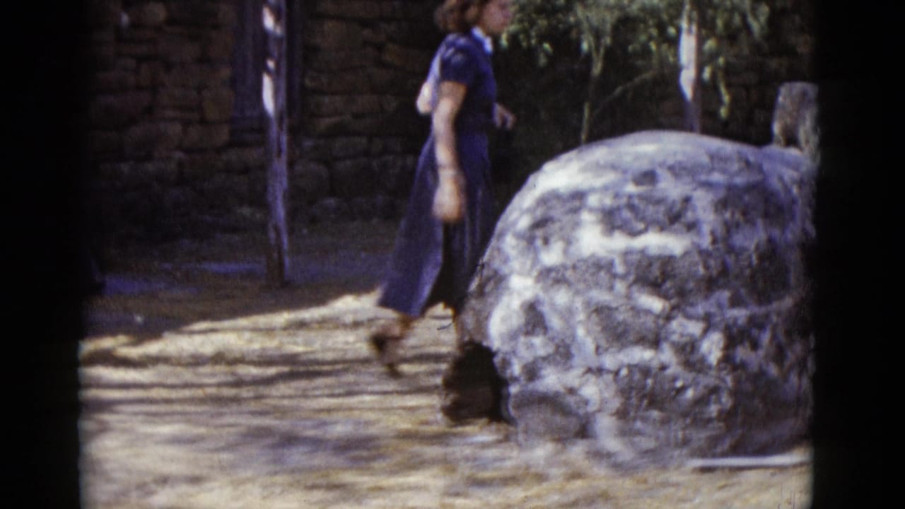 Woman in dark blue 1950s dress walks past a large rounded rough stone structure on a sunny dirt path.