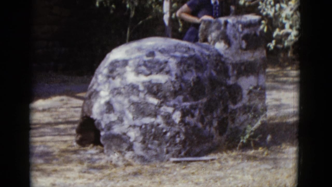 Large rounded rough stone structure dominates the frame; woman in blue dress is partially visible at right edge near a tree.