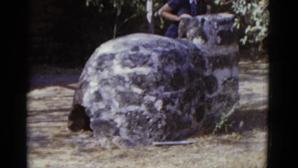 Large weathered rounded stone structure at an outdoor public site; woman in 1950s blue dress exits frame right with a second figure partially visible, 1952.