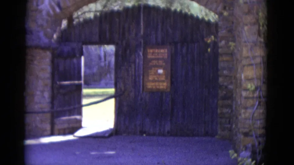 8mm home movie frame from circa 1952 showing a stone archway with open wooden gate, brown entrance sign, and sunlit lawn beyond.