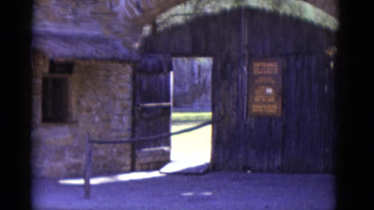Pan left reveals stone building with thatched roof overhang and small window; entrance gate with sign visible at frame edge.