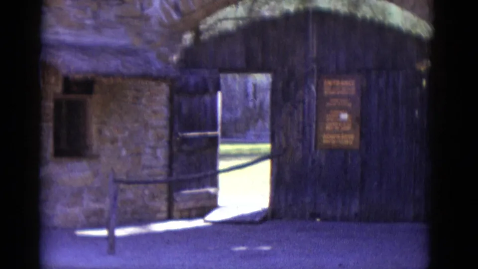 8mm frame circa 1952, pan revealing rustic stone building facade with thatched roof overhang, small window, and partially visible entrance gate at left.