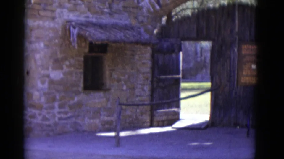 8mm frame circa 1952 showing stone-walled building with thatched overhang, open gate passage, and wooden bench or railing in the foreground.
