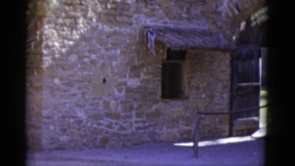 8mm frame circa 1952 showing dressed stone masonry wall with thatched overhang, small window opening, and metal fence post at right.