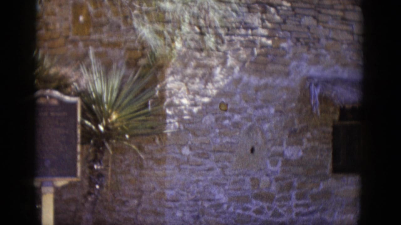 1952 8mm home-movie frame: rough stone wall, two mounted historical marker plaques, and a yucca plant at an apparent Southwestern U.S. landmark.