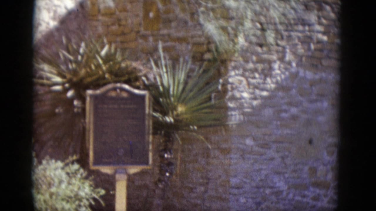 8mm film circa 1952: a mounted historical marker plaque centered before a stone wall with a yucca plant; location appears to be a Southwestern U.S. heritage site.