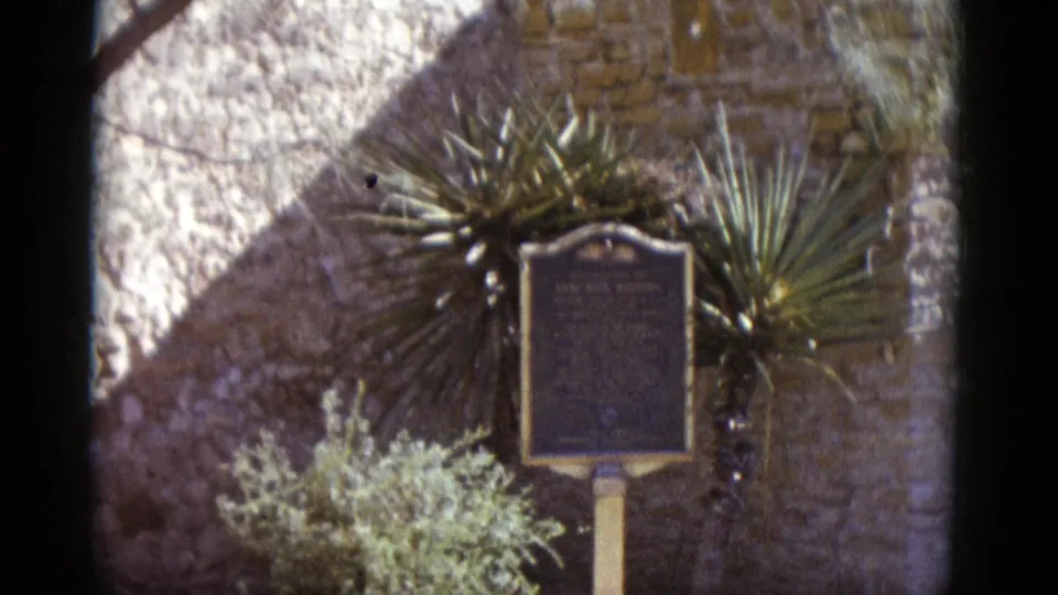 8mm frame circa 1952: historical marker plaque on post framed by a yucca plant against a rough stone wall with strong diagonal sunlight, Southwestern U.S.