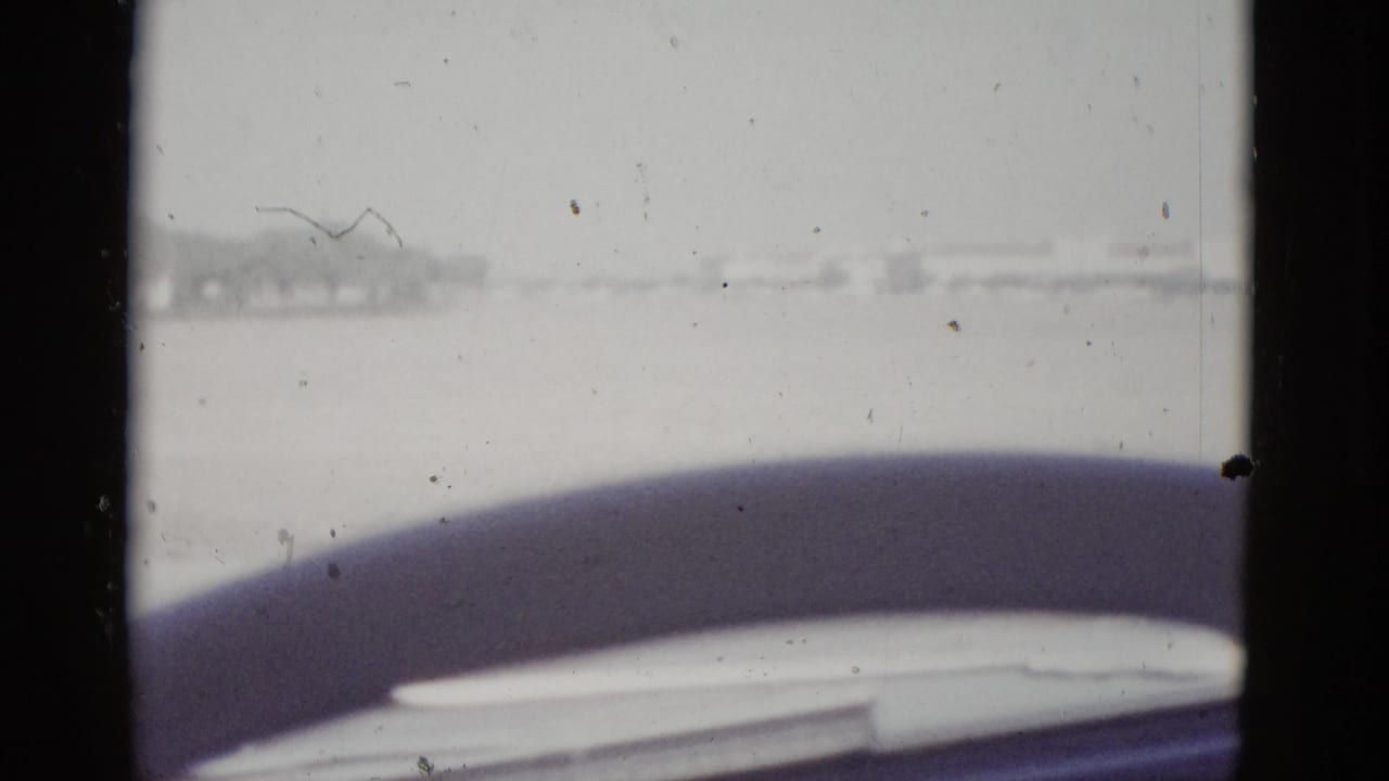 8mm home movie frame circa 1952, shot from inside a moving vehicle in Texas; curved steering wheel fills the foreground while a flat arid landscape and pale sky are visible through the windshield.