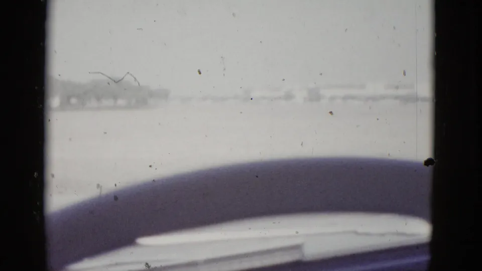 8mm home movie frame circa 1952, shot from inside a moving vehicle in Texas; curved steering wheel fills the foreground while a flat arid landscape and pale sky are visible through the windshield.
