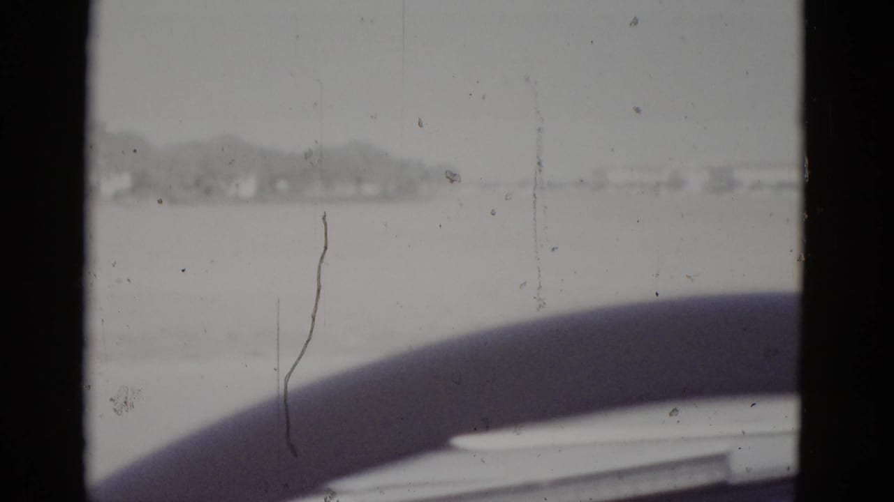Car windshield view with steering wheel in foreground, flat landscape on horizon, and a vertical hairline film scratch visible on the right side of the frame.
