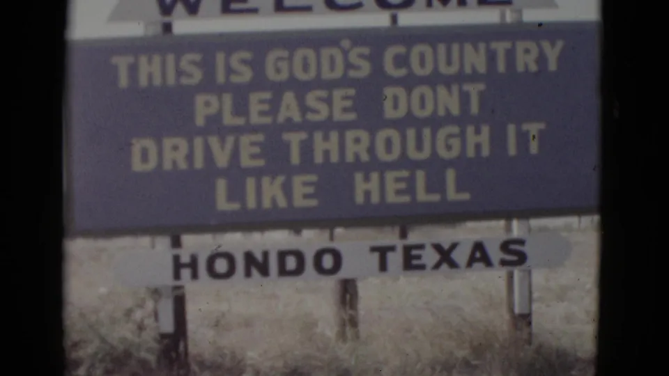 8mm home movie frame circa 1952 showing the Hondo, Texas roadside welcome sign with its civic motto in yellow lettering on a blue background, photographed through a car windshield; sign posts and dry scrub visible at base.