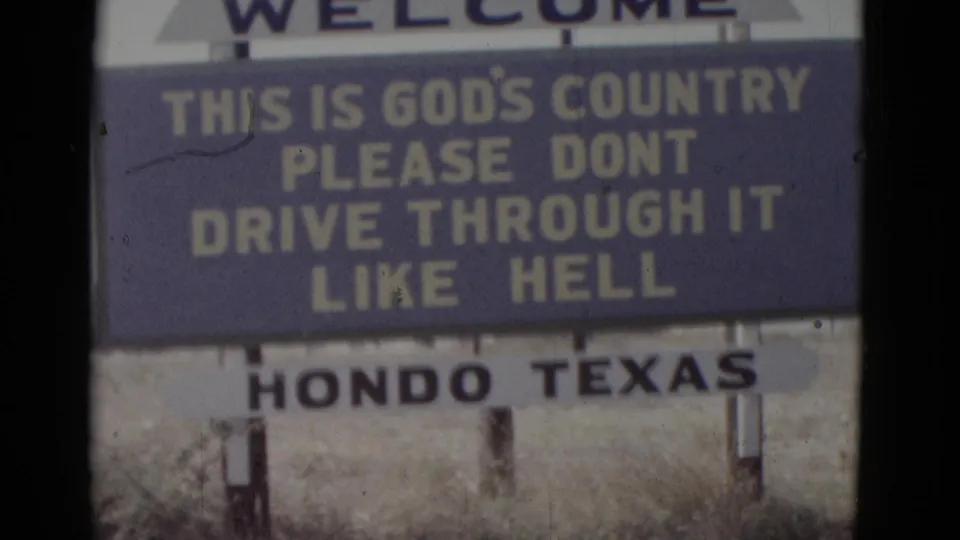 8mm home movie frame circa 1952 showing a close view of the Hondo, Texas civic welcome sign through a car windshield; yellow text on blue background is fully legible, with dry scrub at the sign base.