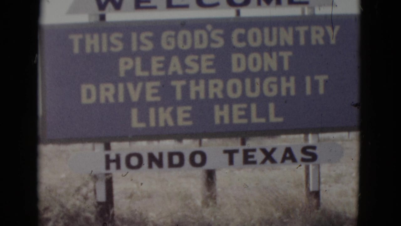 Roadside welcome sign on wooden posts reading THIS IS GOD'S COUNTRY PLEASE DONT DRIVE THROUGH IT LIKE HELL with HONDO TEXAS panel below, in dry scrub grassland.