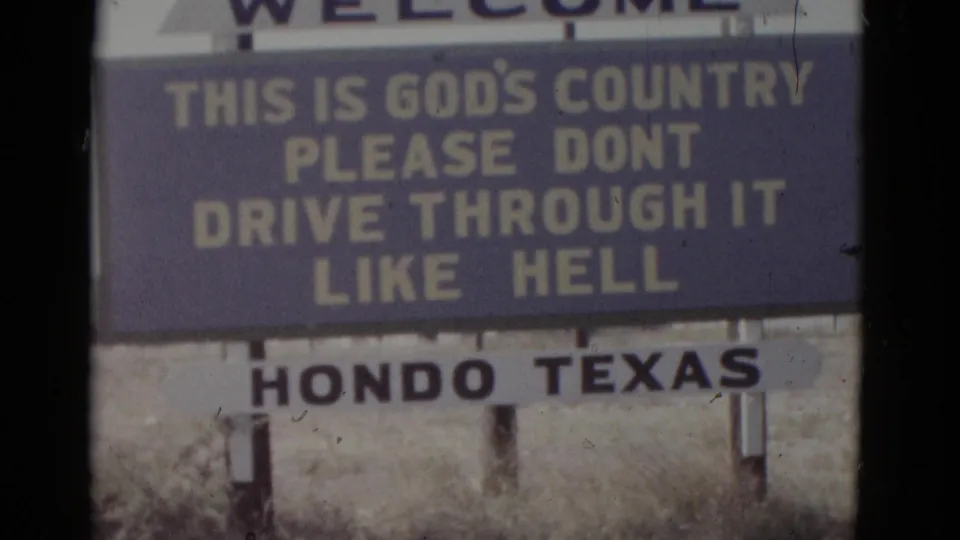 8mm film frame from 1952 showing the Hondo, Texas roadside welcome sign with vernacular humorous text on wooden posts in dry scrub grassland.