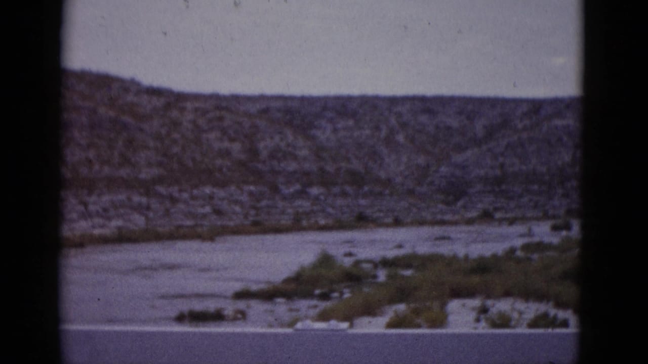Wide shot of a shallow arid river with sandy gravel banks, sparse scrub vegetation, and a rocky ridgeline under an overcast sky.