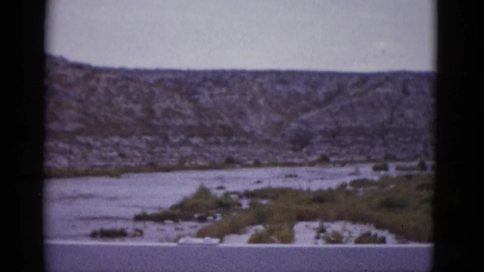 8mm film frame circa 1952 showing the same arid Texas river valley with rocky ridgeline, sparse vegetation, and pale overcast sky.