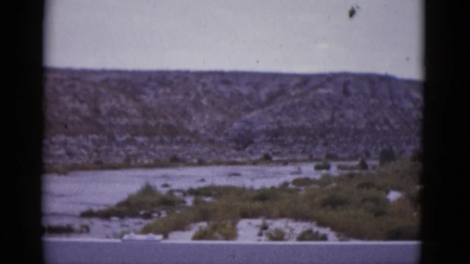 8mm film frame circa 1952 showing a shallow arid river with sandy banks and sparse riparian scrub beneath a rocky Texas hillside.