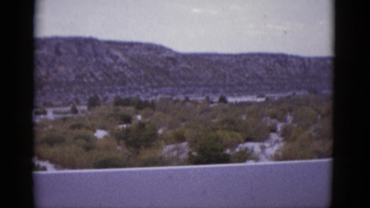 Arid desert valley with a flat-topped mesa on the horizon, scrub vegetation in mid-ground, overcast sky.