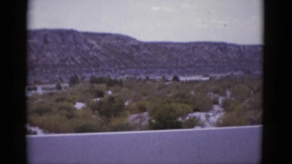 1952 8mm film frame showing a wide Texas desert valley, scrubby low vegetation, and a flat mesa ridge under an overcast sky.