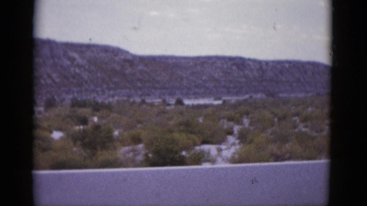Desert mesa panorama at mid-pan, scrub vegetation across valley floor, overcast sky, similar framing to opening frame.