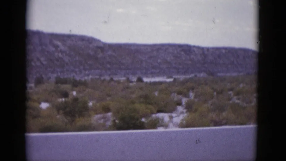 1952 8mm frame of Texas desert terrain with a broad mesa ridge, scrubby valley vegetation, and a hazy pale sky at mid-pan.